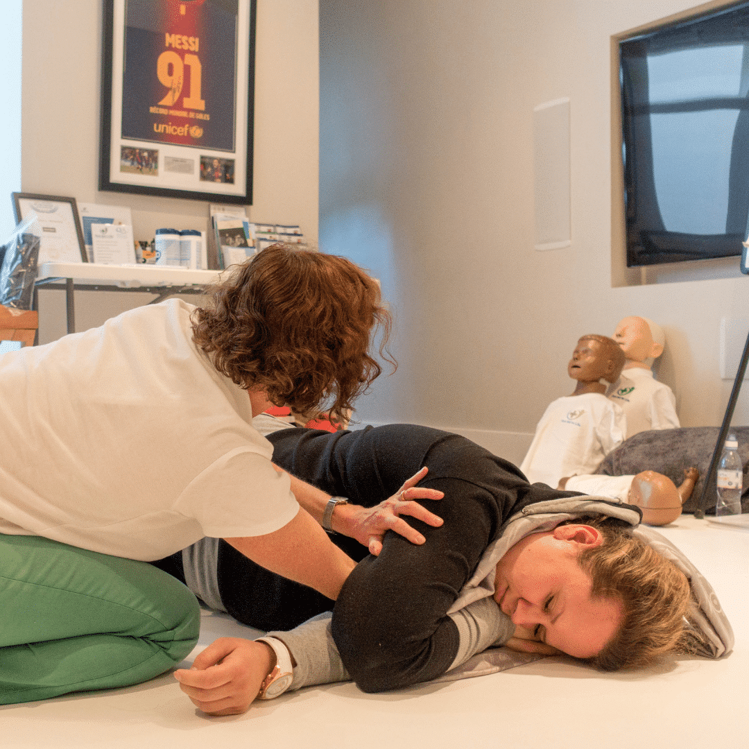 First aid training session showing an instructor placing a woman into the recovery position on the floor, with CPR mannequins visible in the background.