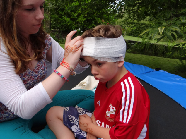 First aid demonstration where a woman is carefully bandaging a young boy’s head injury outdoors, showing how to apply a head bandage in an emergency.